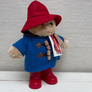May include: A plush bear wearing a bright red felt hat, a blue duffle coat with two wooden toggle closures, and shiny red rain boots. A white and red tag is attached to the coat, featuring the text Paddington and Please look after this bear. Thank you.