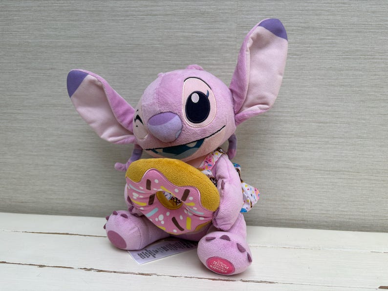 Puede incluir: Un peluche de Angel de Lilo & Stitch, en tonos rosa y morado. El juguete sostiene una rosquilla con glaseado rosa y chispas. El peluche tiene orejas grandes y una expresi&oacute;n traviesa.