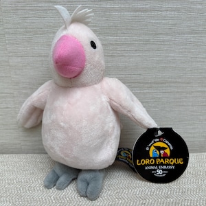 May include: A soft toy parrot, pale pink in colour, with a pink beak and grey feet. It has a white crest and black eyes. A black tag reads "Loro Parque Animal Embassy".