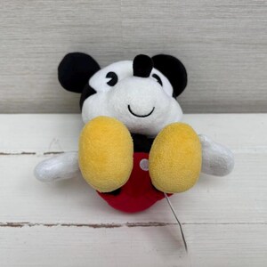 Op de afbeelding: Een pluche Mickey Mouse-speelgoed met een zwart-witte kop, zwarte oren en gele voeten. Het speelgoed heeft een rode onderkant en witte armen. Er is een klein zilveren label bevestigd.