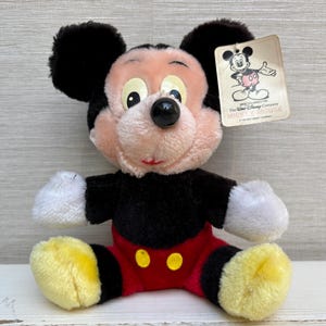 Puede incluir: Un peluche vintage de Mickey Mouse. El peluche presenta un cuerpo negro, pantalones cortos rojos con botones amarillos y pies amarillos. Tiene una cara rosa, orejas negras y una etiqueta con una ilustraci&oacute;n de Mickey Mouse.
