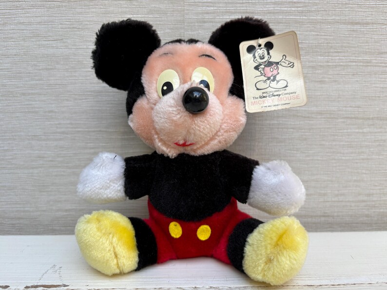 Puede incluir: Un peluche vintage de Mickey Mouse. El peluche presenta un cuerpo negro, pantalones cortos rojos con botones amarillos y pies amarillos. Tiene una cara rosa, orejas negras y una etiqueta con una ilustraci&oacute;n de Mickey Mouse.