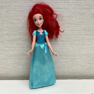 Op de afbeelding: Pop van een personage met lang, levendig rood haar, gekleed in een teal jurk met een sprankelend lijfje. De pop heeft een glimlachende uitdrukking en poseert tegen een neutrale achtergrond.