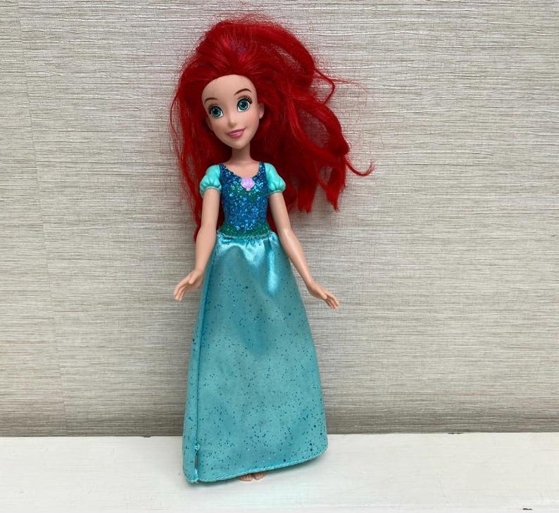 Op de afbeelding: Pop van een personage met lang, levendig rood haar, gekleed in een teal jurk met een sprankelend lijfje. De pop heeft een glimlachende uitdrukking en poseert tegen een neutrale achtergrond.