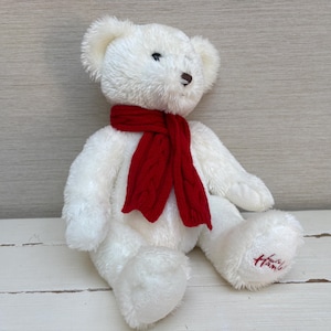 May include: A white plush teddy bear with a red scarf. The bear has a brown nose and black eyes. The bear is sitting on a white surface. The bear's foot has the word "Harrods" embroidered on it.