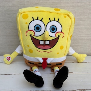 May include: A plush SpongeBob SquarePants toy featuring a bright yellow rectangular body with brown trousers, a white shirt, and a red tie. The character has large blue eyes, a wide smiling mouth with two teeth, and a Build-A-Bear Workshop logo on one hand.