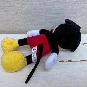 May include: A plush Mickey Mouse toy, lying on its side. Mickey is wearing red shorts, yellow shoes, white gloves, and a black body and head. The Disney tag is visible. The toy is on a white wooden surface.