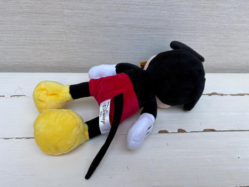 May include: A plush Mickey Mouse toy, lying on its side. Mickey is wearing red shorts, yellow shoes, white gloves, and a black body and head. The Disney tag is visible. The toy is on a white wooden surface.