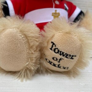 May include: A plush teddy bear with tan fur and a red and white uniform. The bear's paws are visible, with the words "Tower of London" printed on one paw in black.