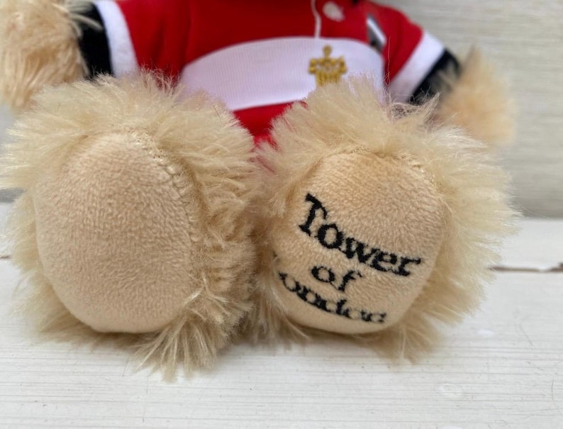 May include: A plush teddy bear with tan fur and a red and white uniform. The bear's paws are visible, with the words "Tower of London" printed on one paw in black.