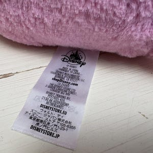 May include: Close-up of a soft, light purple plush toy. A white tag with the Disney logo and text is visible, including "MADE IN CHINA" and "shopDisney.com". The tag also includes the address for The Disney Store Ltd in London.