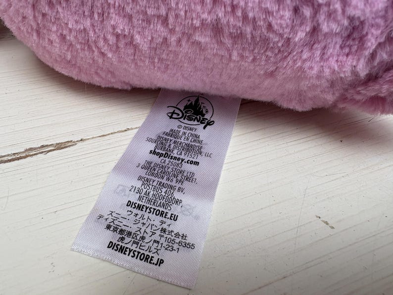 May include: Close-up of a soft, light purple plush toy. A white tag with the Disney logo and text is visible, including "MADE IN CHINA" and "shopDisney.com". The tag also includes the address for The Disney Store Ltd in London.