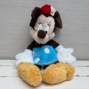 May include: A plush Minnie Mouse doll with a red bow and white flower on her head. She has a light brown face, dark brown ears, and a blue dress with white polka dots. Her hands and feet are white and yellow.