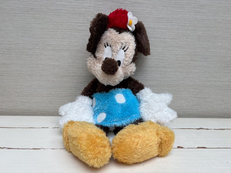 May include: A plush Minnie Mouse doll with a red bow and white flower on her head. She has a light brown face, dark brown ears, and a blue dress with white polka dots. Her hands and feet are white and yellow.