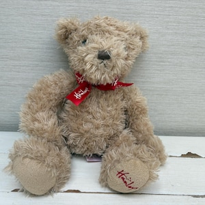 May include: A light brown plush teddy bear with shaggy fur, featuring a dark brown stitched nose and eyes. A red ribbon is tied around its neck with the white text Hamleys printed on it. The left foot pad features the red embroidered text Hamleys.