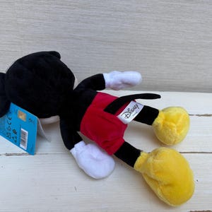 May include: A plush Mickey Mouse toy, lying on its side. The toy features a black head and body, red shorts, yellow feet, and white gloves. A Disney tag is attached. The toy is on a light-colored wooden surface.