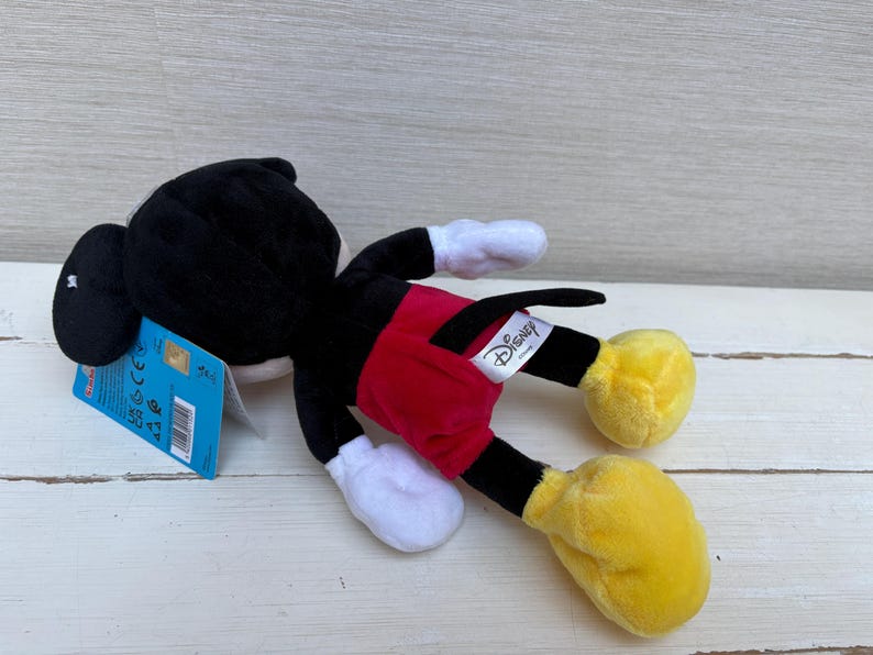 May include: A plush Mickey Mouse toy, lying on its side. The toy features a black head and body, red shorts, yellow feet, and white gloves. A Disney tag is attached. The toy is on a light-colored wooden surface.
