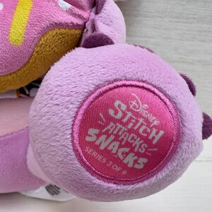 Puede incluir: Primer plano de un peluche con una rosquilla rosa y un pie lavanda con un emblema circular. El emblema dice "Disney Stitch Attacks Snacks Serie 3 de 9" en texto rosa.