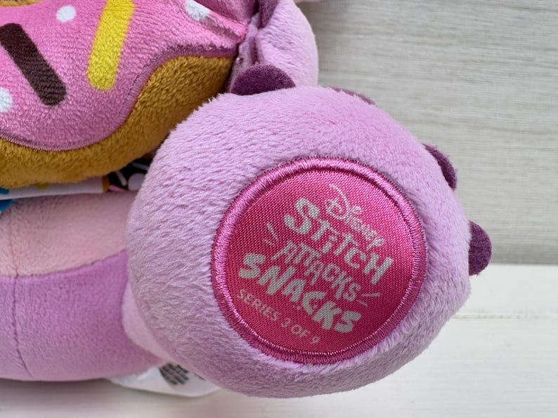 Puede incluir: Primer plano de un peluche con una rosquilla rosa y un pie lavanda con un emblema circular. El emblema dice "Disney Stitch Attacks Snacks Serie 3 de 9" en texto rosa.