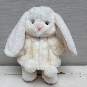 Könnte beinhalten: Ein cremefarbenes Plüschhasen-Spielzeug mit langen, hängenden Ohren und einer kleinen rosa Nase. Der Hase trägt eine passende cremefarbene Kapuzenjacke aus Kunstfell mit zwei herabhängenden geflochtenen Kordeln und Bommeln.