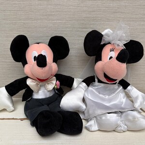 May include: Two plush Mickey and Minnie Mouse dolls dressed for a wedding. Mickey wears a tuxedo with a bow tie and Minnie wears a white wedding dress and veil. The dolls have black ears, pink faces, and white gloves.