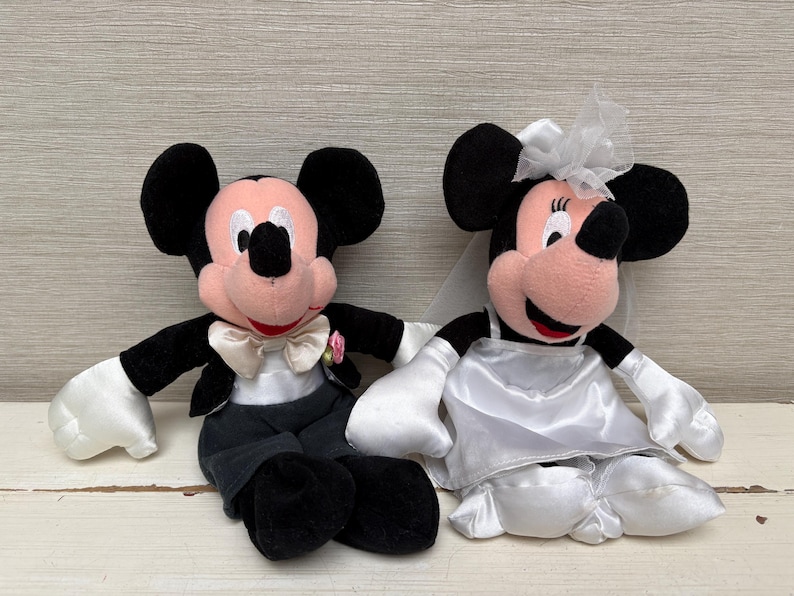 May include: Two plush Mickey and Minnie Mouse dolls dressed for a wedding. Mickey wears a tuxedo with a bow tie and Minnie wears a white wedding dress and veil. The dolls have black ears, pink faces, and white gloves.