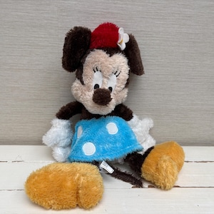May include: A plush Minnie Mouse doll with brown ears, a red bow, and a blue dress with white polka dots. The doll has white gloves and yellow shoes. The toy is sitting on a white surface.