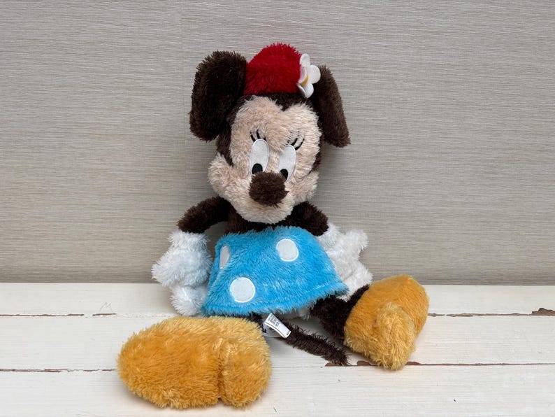 May include: A plush Minnie Mouse doll with brown ears, a red bow, and a blue dress with white polka dots. The doll has white gloves and yellow shoes. The toy is sitting on a white surface.