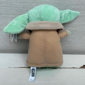 Peut inclure: Une peluche d'un personnage avec de grandes oreilles et une robe beige. Le jouet a une t&ecirc;te et des oreilles vert clair, avec un cou et un corps beiges. Le jouet est pos&eacute; sur une surface en bois blanc. Une &eacute;tiquette Star Wars est attach&eacute;e.