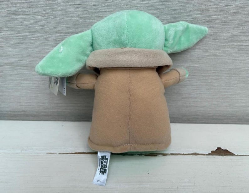 Peut inclure: Une peluche d'un personnage avec de grandes oreilles et une robe beige. Le jouet a une t&ecirc;te et des oreilles vert clair, avec un cou et un corps beiges. Le jouet est pos&eacute; sur une surface en bois blanc. Une &eacute;tiquette Star Wars est attach&eacute;e.