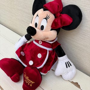 May include: A Minnie Mouse soft toy, seated and wearing a red dress with white trim and polka dots, a matching bow, and black ears. Her white gloved hands and red shoes are visible. The text "Disney Store 2014" is embroidered on her shoe.