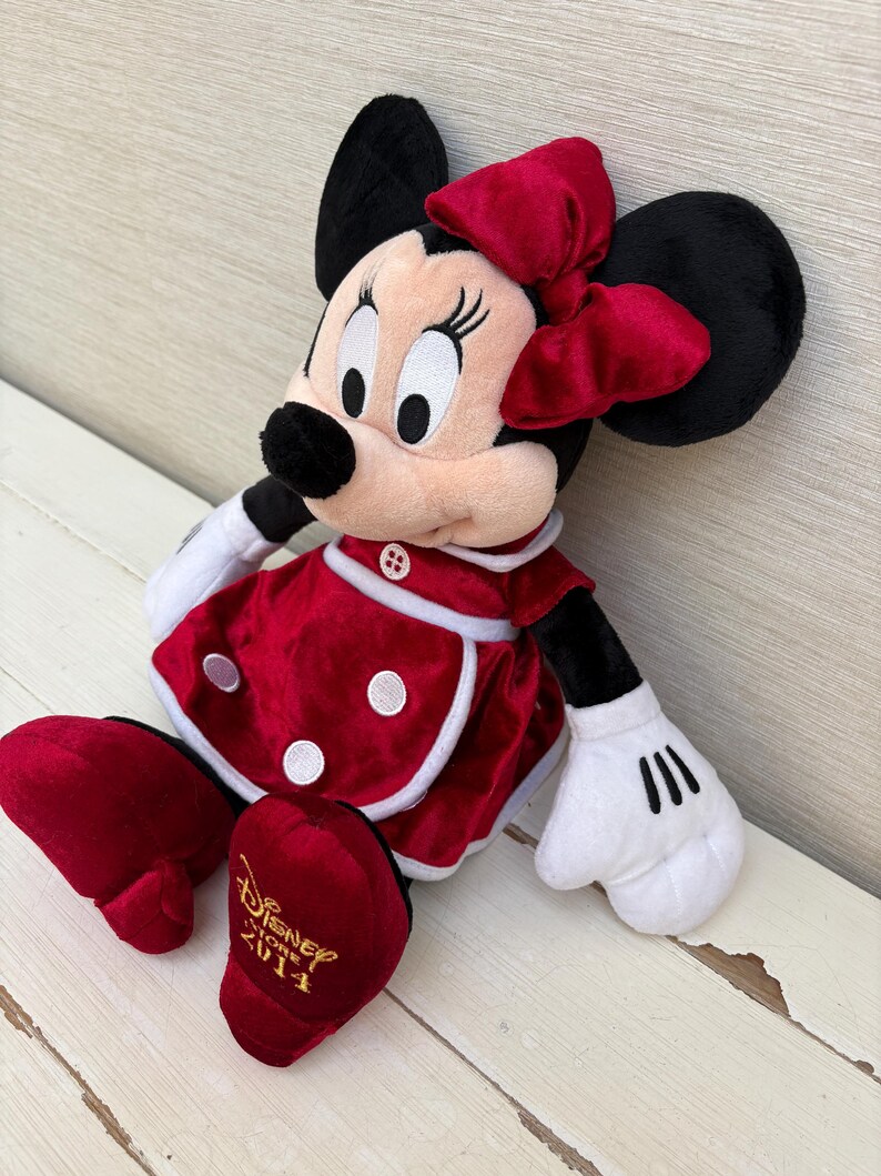 May include: A Minnie Mouse soft toy, seated and wearing a red dress with white trim and polka dots, a matching bow, and black ears. Her white gloved hands and red shoes are visible. The text "Disney Store 2014" is embroidered on her shoe.
