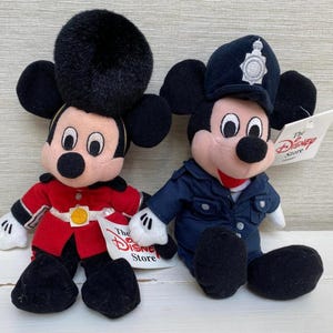 May include: Two Mickey Mouse soft toys. One is dressed in a red military uniform with a black hat, the other in a blue police uniform with a badge. Both have black ears, a pink face, and a black nose. Each has a Disney Store tag.