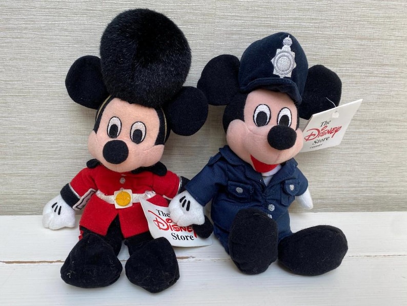 May include: Two Mickey Mouse soft toys. One is dressed in a red military uniform with a black hat, the other in a blue police uniform with a badge. Both have black ears, a pink face, and a black nose. Each has a Disney Store tag.