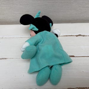 Może przedstawiać: Pluszowa lalka Minnie Mouse, widoczna od tyłu, w turkusowej sukience i kokardzie. Lalka ma czarne uszy i r&oacute;żową twarz. Jej dłonie są białe, a stopy turkusowe. Lalka leży na białej drewnianej powierzchni.