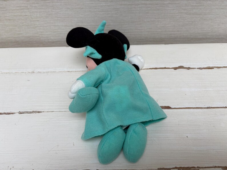 Może przedstawiać: Pluszowa lalka Minnie Mouse, widoczna od tyłu, w turkusowej sukience i kokardzie. Lalka ma czarne uszy i r&oacute;żową twarz. Jej dłonie są białe, a stopy turkusowe. Lalka leży na białej drewnianej powierzchni.