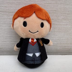 May include: A plush doll featuring a character with bright orange hair, black embroidered eyes, and a small smile. The doll wears a black robe over a gray vest, a white shirt, and a red and gold striped tie, with a gold crest on the left chest.