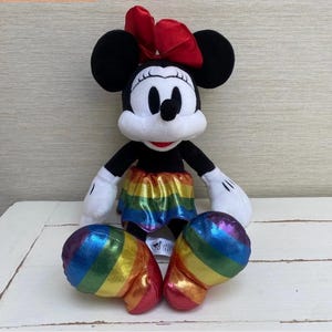 May include: A Minnie Mouse soft toy featuring a red bow, black ears, and a rainbow skirt. The toy has white gloves and rainbow-coloured shoes. The plush is positioned on a white surface.