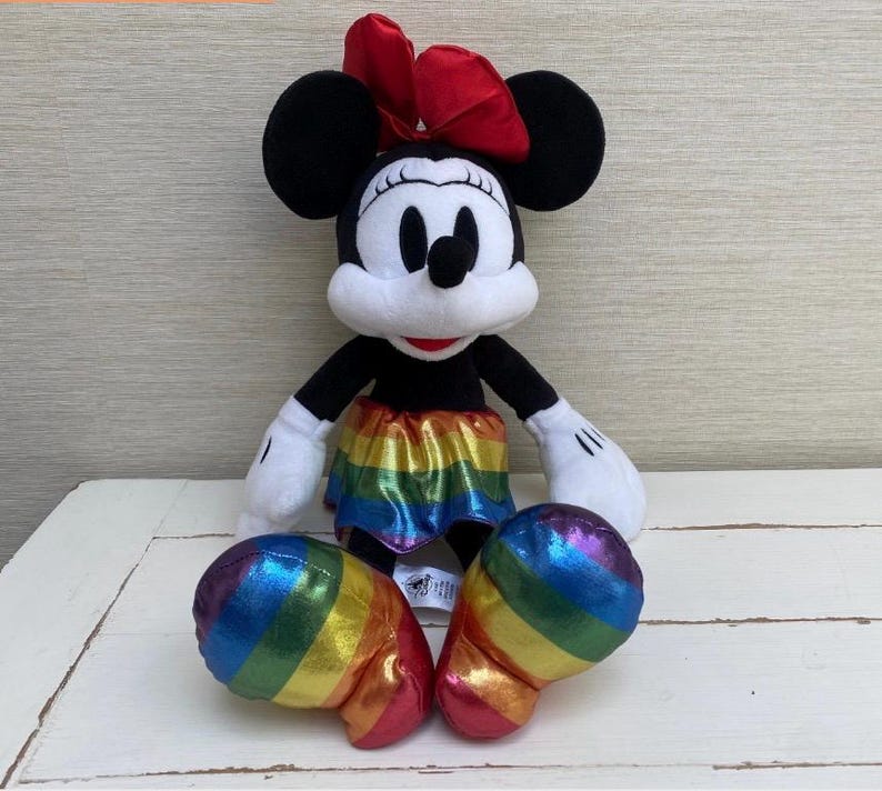 May include: A Minnie Mouse soft toy featuring a red bow, black ears, and a rainbow skirt. The toy has white gloves and rainbow-coloured shoes. The plush is positioned on a white surface.