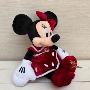 May include: A plush Minnie Mouse doll, with black ears, a red bow, and a red dress with white polka dots. The doll has white gloves and red shoes. The Disney Store 2014 logo is on the shoe.