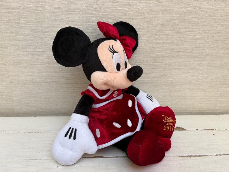 May include: A plush Minnie Mouse doll, with black ears, a red bow, and a red dress with white polka dots. The doll has white gloves and red shoes. The Disney Store 2014 logo is on the shoe.