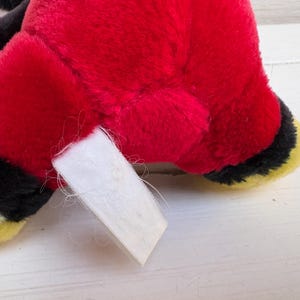 Puede incluir: Primer plano del cuerpo rojo de un peluche, con detalles en negro y amarillo y una etiqueta blanca. El peluche tiene un pelaje suave y texturizado. La etiqueta est&aacute; en blanco y est&aacute; sujeta al peluche.