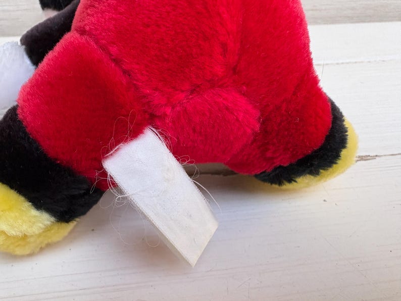 Puede incluir: Primer plano del cuerpo rojo de un peluche, con detalles en negro y amarillo y una etiqueta blanca. El peluche tiene un pelaje suave y texturizado. La etiqueta est&aacute; en blanco y est&aacute; sujeta al peluche.