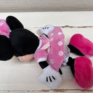 May include: A plush Minnie Mouse toy, featuring black ears, a pink and white polka dot bow, and pink shoes. The toy is lying on a white wooden surface.