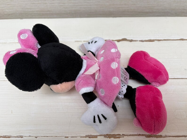 May include: A plush Minnie Mouse toy, featuring black ears, a pink and white polka dot bow, and pink shoes. The toy is lying on a white wooden surface.