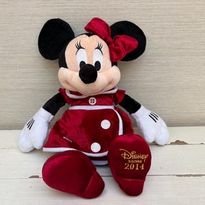 May include: A plush Minnie Mouse doll in a red dress with white accents, a matching bow, and black ears. The doll has white gloves and a black nose. The right foot has the text "Disney Store 2014" in gold.