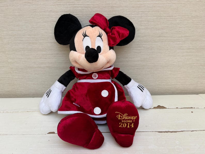 May include: A plush Minnie Mouse doll in a red dress with white accents, a matching bow, and black ears. The doll has white gloves and a black nose. The right foot has the text "Disney Store 2014" in gold.