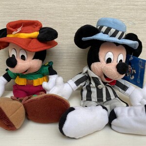 May include: Two Mickey Mouse soft toys. One wears a red hat with a yellow band and a green shirt. The other Mickey Mouse wears a light blue hat with black and white stripes and a black and white striped shirt. Both have white gloves and black ears.