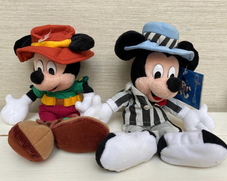 May include: Two Mickey Mouse soft toys. One wears a red hat with a yellow band and a green shirt. The other Mickey Mouse wears a light blue hat with black and white stripes and a black and white striped shirt. Both have white gloves and black ears.