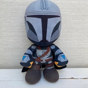May include: A plush toy depicting a character from a popular science fiction series. The figure has a gray helmet and a detailed outfit with brown and black accents. The toy has a black cape and brown boots.